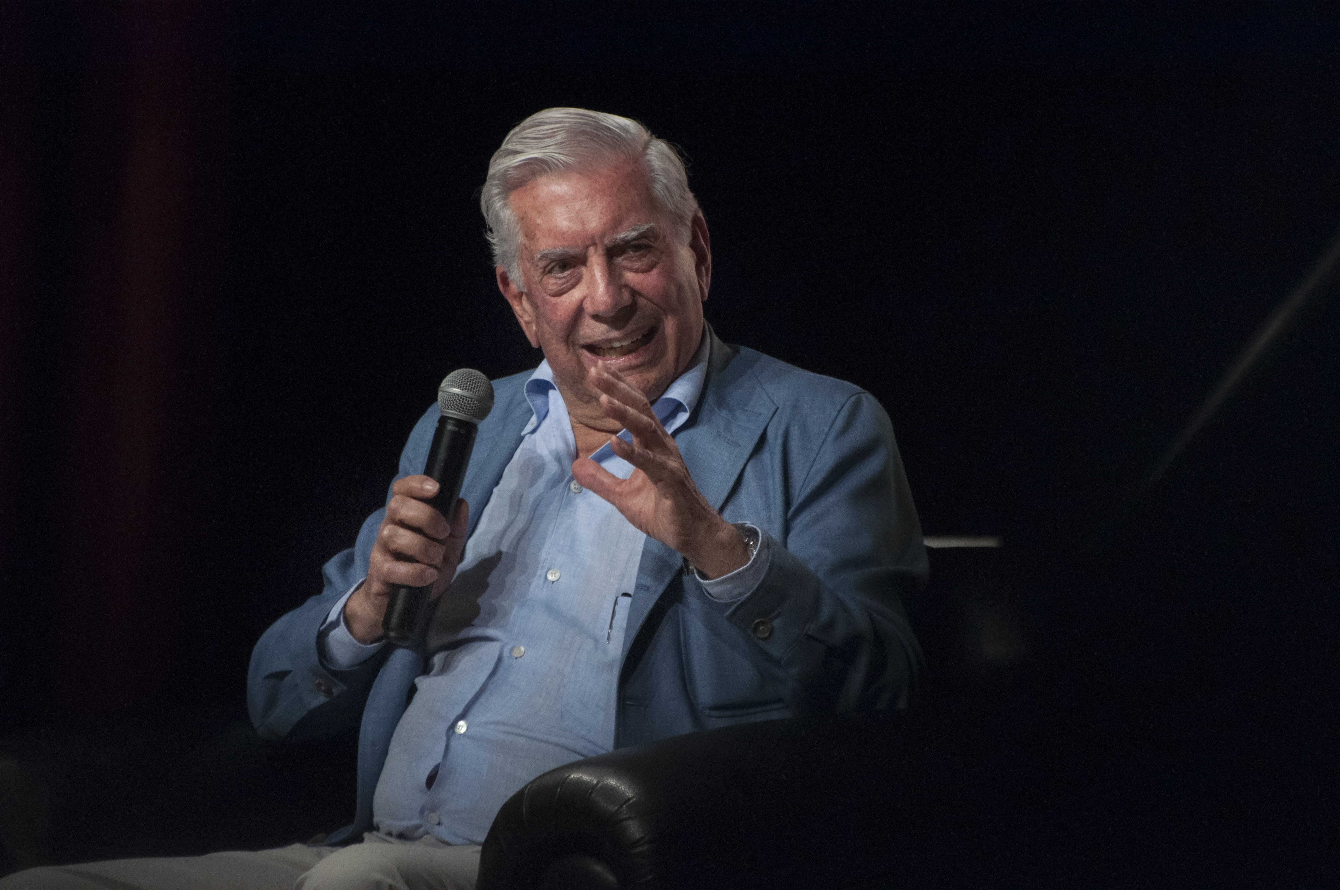 Foto del escritor Mario Vargas Llosa sentado en un butacón, en actitud de conversación, con expresión sonriente mientras sostiene en una mano un micrófono.